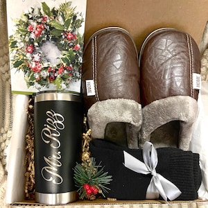 Pode incluir: Uma caixa de presente cheia de uma caneca de viagem preta personalizada com o texto "Mr. Riggs", um par de pantuflas de couro sint&eacute;tico marrom com a palavra "HOME" nelas, um par de meias pretas com uma fita prateada e um cart&atilde;o de Natal com uma coroa.