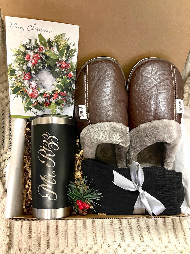 Pode incluir: Uma caixa de presente cheia de uma caneca de viagem preta personalizada com o texto "Mr. Riggs", um par de pantuflas de couro sint&eacute;tico marrom com a palavra "HOME" nelas, um par de meias pretas com uma fita prateada e um cart&atilde;o de Natal com uma coroa.