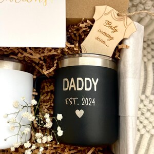 Puede incluir: Una caja de regalo que contiene una taza negra con la inscripci&oacute;n "DADDY EST. 2024", una taza blanca, una etiqueta de madera con forma de body de beb&eacute; que dice "Baby coming soon..." y una tarjeta que dice "&iexcl;Enhorabuena!". La caja est&aacute; llena de papel marr&oacute;n y flores blancas.