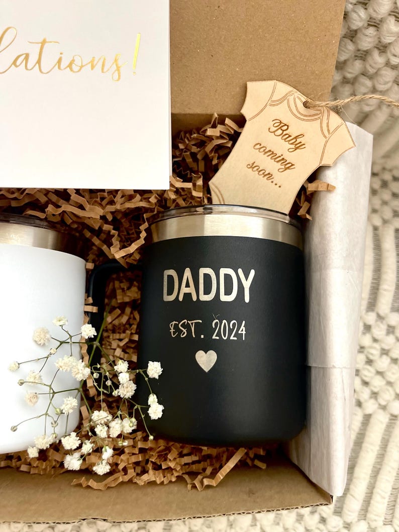 Puede incluir: Una caja de regalo que contiene una taza negra con la inscripci&oacute;n "DADDY EST. 2024", una taza blanca, una etiqueta de madera con forma de body de beb&eacute; que dice "Baby coming soon..." y una tarjeta que dice "&iexcl;Enhorabuena!". La caja est&aacute; llena de papel marr&oacute;n y flores blancas.