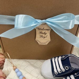 Może przedstawiać: Brązowe pudełko prezentowe zawiązane jasnoniebieską satynową wstążką i drewnianą etykietą z napisem "Welcome Baby Boy!". Zawiera granatowe buciki dla dziecka z białymi sznurowadłami oraz niebieski i drewniany klips do smoczka.