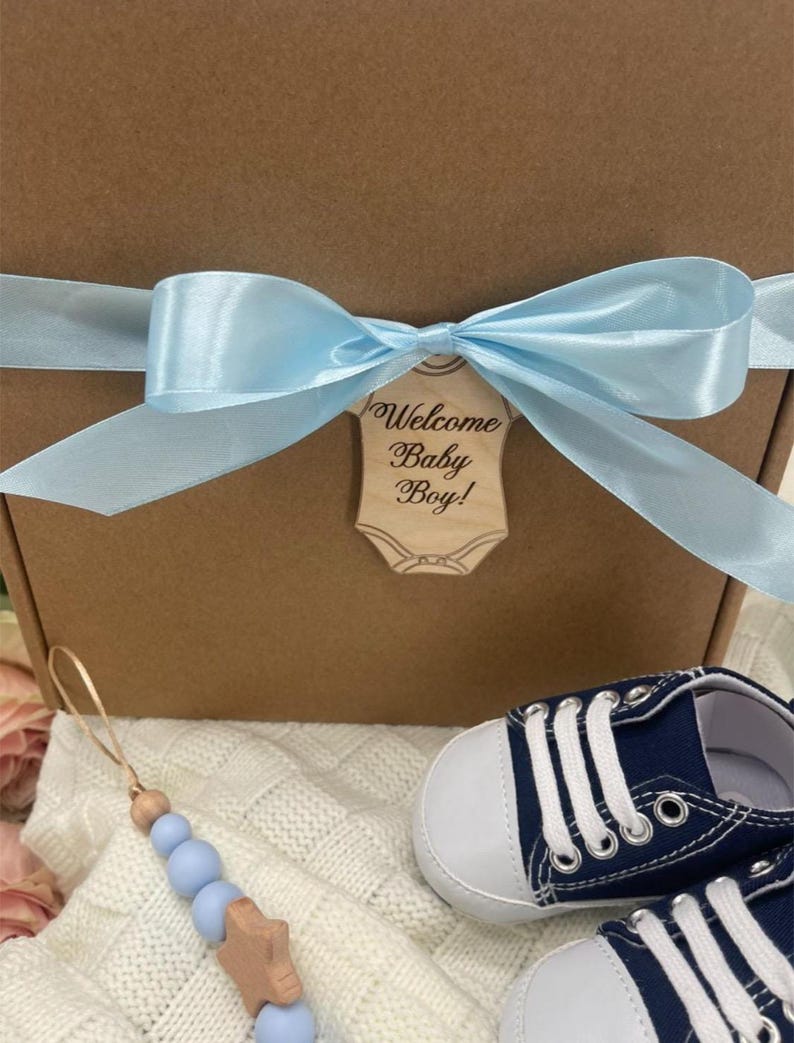 Może przedstawiać: Brązowe pudełko prezentowe zawiązane jasnoniebieską satynową wstążką i drewnianą etykietą z napisem "Welcome Baby Boy!". Zawiera granatowe buciki dla dziecka z białymi sznurowadłami oraz niebieski i drewniany klips do smoczka.