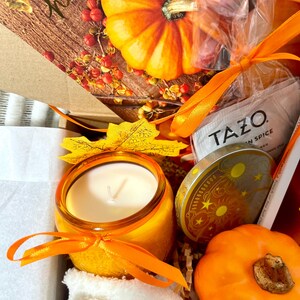 May include: A gift box filled with fall-themed items, including a candle, a pumpkin, a tea bag, and a small decorative tin. The box is lined with white tissue paper and has a card that says "Happy Fall".