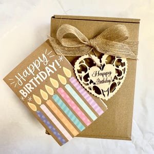 May include: A brown gift box tied with a burlap bow, accompanied by a birthday card and a wooden heart-shaped tag. The card reads "Happy BIRTHDAY!" and features colorful candles. The wooden tag also says "Happy Birthday!"