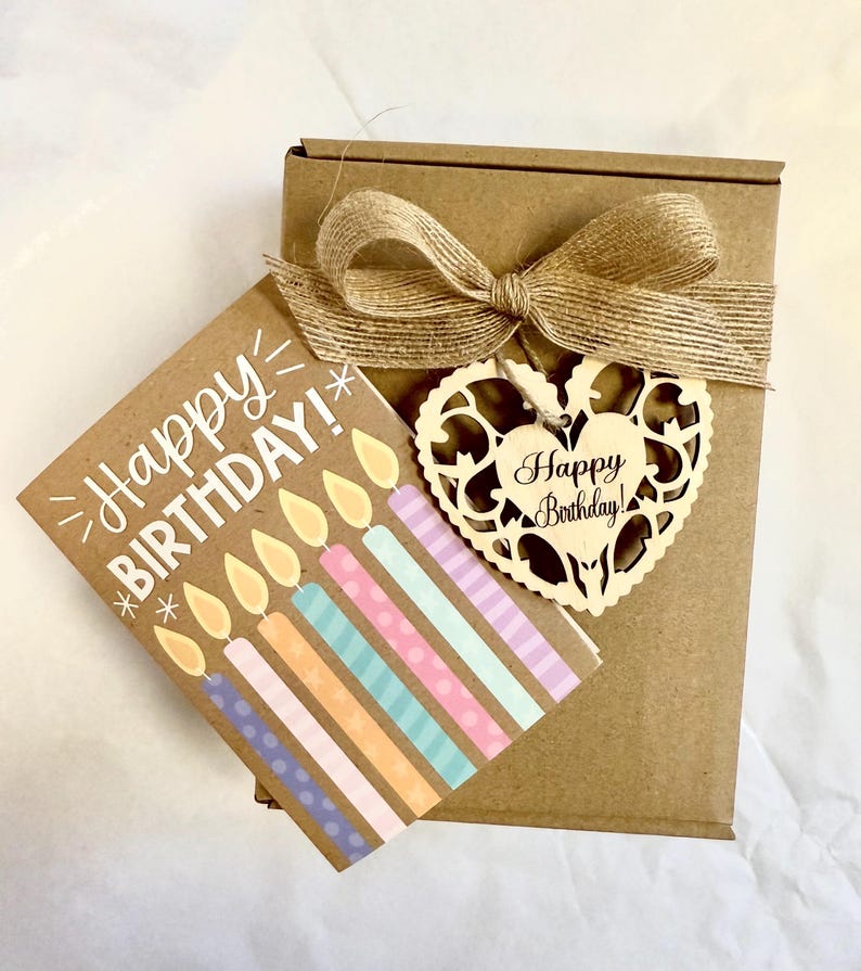 May include: A brown gift box tied with a burlap bow, accompanied by a birthday card and a wooden heart-shaped tag. The card reads "Happy BIRTHDAY!" and features colorful candles. The wooden tag also says "Happy Birthday!"