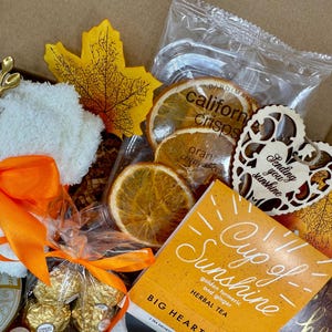 May include: A gift box filled with a variety of items. Includes dried orange slices, a wooden heart with the text "Sending you sunshine", a tea packet, a gold spoon, and a white washcloth with an orange bow.