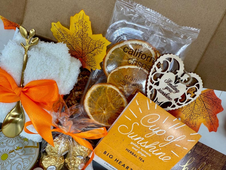 May include: A gift box filled with a variety of items. Includes dried orange slices, a wooden heart with the text "Sending you sunshine", a tea packet, a gold spoon, and a white washcloth with an orange bow.