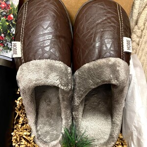 Pu&ograve; includere: Un paio di pantofole in similpelle marrone con fodera in pile grigio. Le pantofole hanno un'etichetta "HOME" su ogni lato.