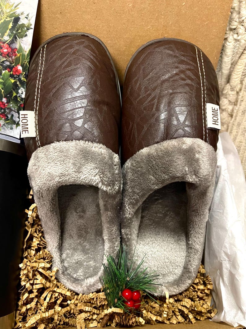 Pu&ograve; includere: Un paio di pantofole in similpelle marrone con fodera in pile grigio. Le pantofole hanno un'etichetta "HOME" su ogni lato.