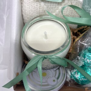 May include: A gift basket with a white knitted blanket, a candle in a clear glass jar with a white wax, a green ribbon, and a round tin with a white and gold design.