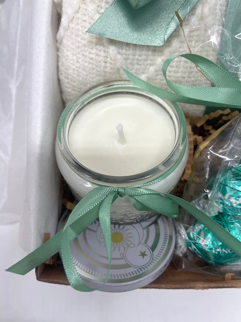 May include: A gift basket with a white knitted blanket, a candle in a clear glass jar with a white wax, a green ribbon, and a round tin with a white and gold design.