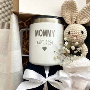 May include: A gift box filled with a white mug with the text "Mommy Est. 2024" and a heart, a white crocheted bunny, a pair of white socks, and a black fabric with a white ribbon.