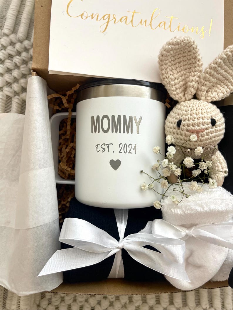 May include: A gift box filled with a white mug with the text "Mommy Est. 2024" and a heart, a white crocheted bunny, a pair of white socks, and a black fabric with a white ribbon.