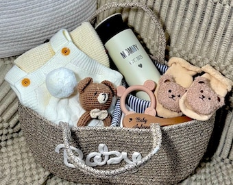 May include: A brown wicker basket filled with baby gifts, including a knitted white onesie, a brown crocheted teddy bear, a wooden spoon with the text "Baby Anderson", a beige and brown baby sock set, a beige and brown teething ring, and a white thermos with the text "Mommy Est. 2023".