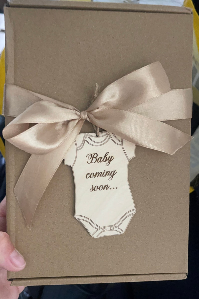 May include: A brown cardboard box with a beige satin ribbon tied in a bow. A wooden cutout of a onesie with the text "Baby coming soon..." is attached to the ribbon.