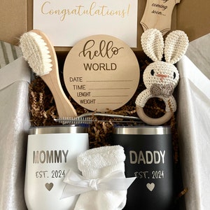 May include: A gift box filled with baby shower gifts, including a wooden sign that says "hello world", a white baby brush, a crocheted bunny rattle, two stainless steel tumblers with the words "mommy est. 2024" and "daddy est. 2024", a pair of white baby socks, and a card that says "congratulations!" and "baby coming soon..."