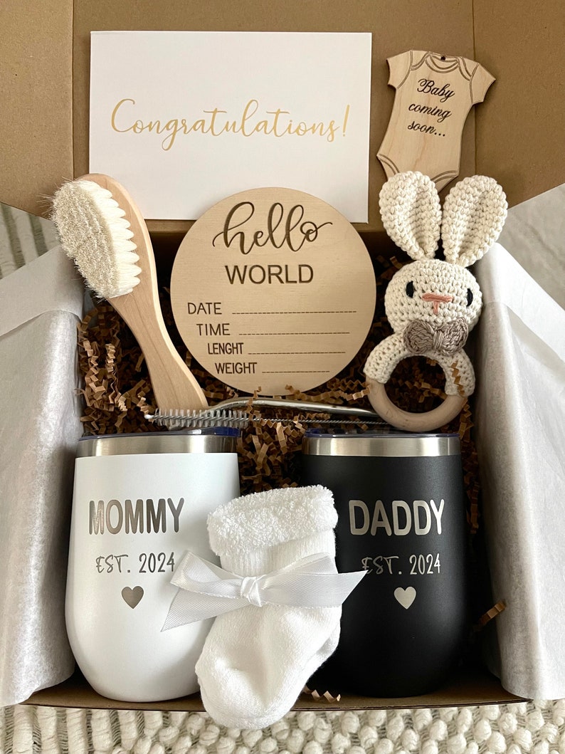 May include: A gift box filled with baby shower gifts, including a wooden sign that says "hello world", a white baby brush, a crocheted bunny rattle, two stainless steel tumblers with the words "mommy est. 2024" and "daddy est. 2024", a pair of white baby socks, and a card that says "congratulations!" and "baby coming soon..."