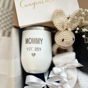 May include: A gift box filled with a white stainless steel tumbler with the text "Mommy Est. 2024" and a heart, a white crocheted bunny, a pair of white socks, and a black and white ribbon.