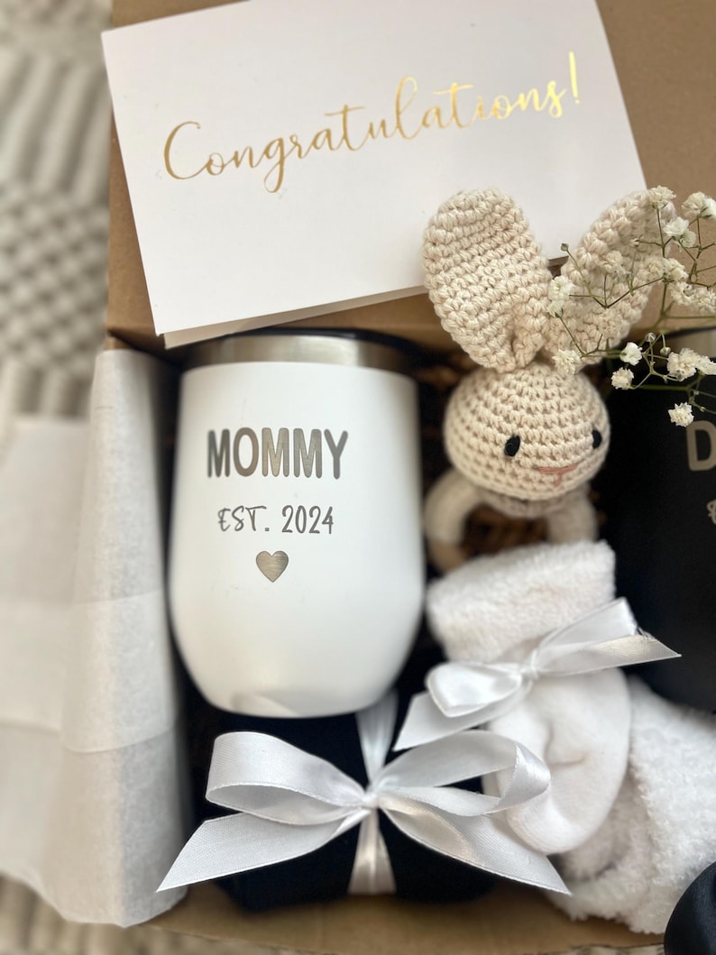May include: A gift box filled with a white stainless steel tumbler with the text "Mommy Est. 2024" and a heart, a white crocheted bunny, a pair of white socks, and a black and white ribbon.