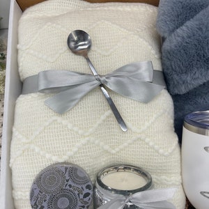 Op de afbeelding: Een cadeaupakket met een wit gebreid deken, een zilveren lepel, een zilveren kaars met witte was, een zilveren pot met een bloemenpatroon en een witte mok met een zilveren deksel.