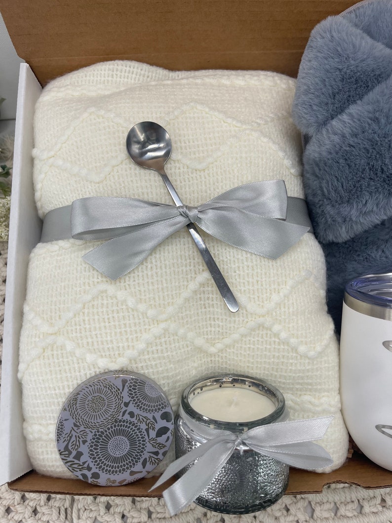 Op de afbeelding: Een cadeaupakket met een wit gebreid deken, een zilveren lepel, een zilveren kaars met witte was, een zilveren pot met een bloemenpatroon en een witte mok met een zilveren deksel.