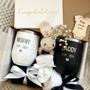 May include: A gift box filled with a variety of items for a new parent. The box includes two black and white mugs with the words "Mommy Est. 2024" and "Daddy Est. 2024" on them, a white crocheted bunny, a wooden ornament that says "Baby coming soon...", and two white socks with black bows.