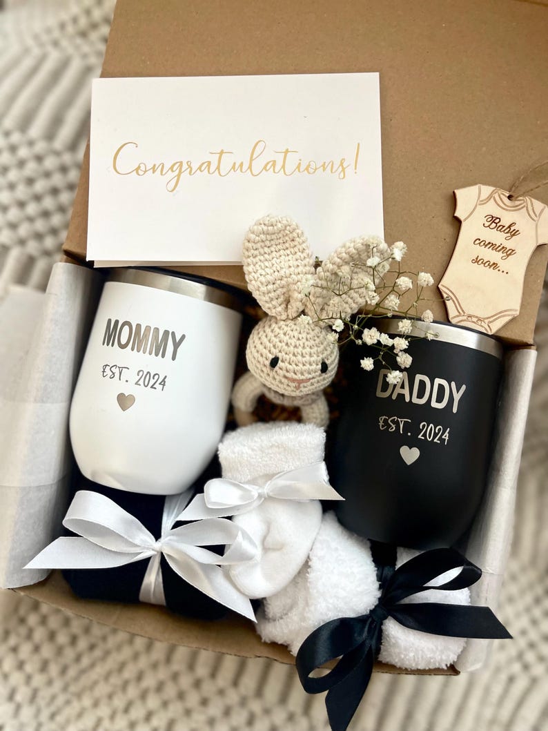 May include: A gift box filled with a variety of items for a new parent. The box includes two black and white mugs with the words "Mommy Est. 2024" and "Daddy Est. 2024" on them, a white crocheted bunny, a wooden ornament that says "Baby coming soon...", and two white socks with black bows.