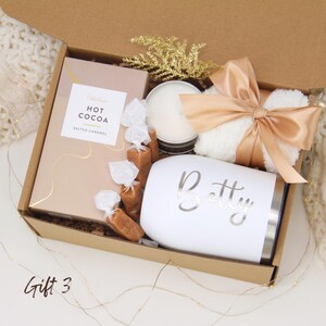 Puede incluir: Una caja de regalo llena con un vaso blanco con "Betty" impreso, una vela, una caja de chocolate caliente, caramelos envueltos y un art&iacute;culo blanco esponjoso atado con una cinta color melocot&oacute;n. La caja es marr&oacute;n y tiene una ramita dorada brillante.