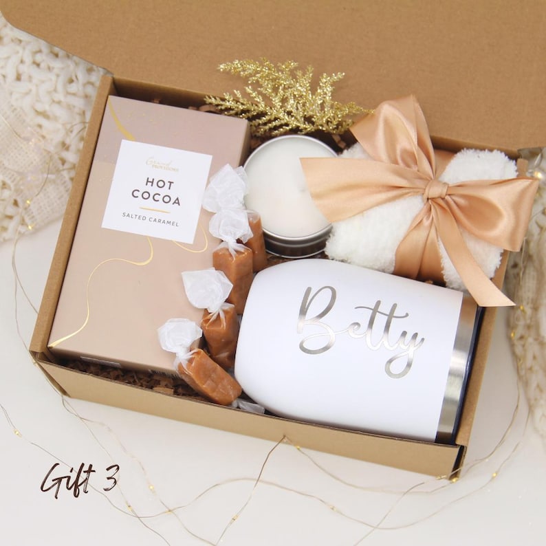 Puede incluir: Una caja de regalo llena con un vaso blanco con "Betty" impreso, una vela, una caja de chocolate caliente, caramelos envueltos y un art&iacute;culo blanco esponjoso atado con una cinta color melocot&oacute;n. La caja es marr&oacute;n y tiene una ramita dorada brillante.