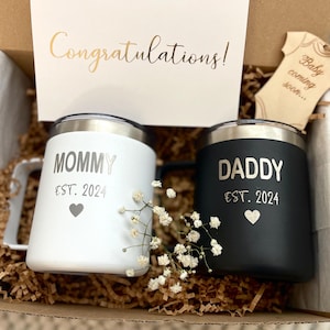 Puede incluir: Una caja de regalo con dos tazas aisladas, una blanca y otra negra, con bordes plateados. La taza blanca dice "MOMMY EST. 2024" y la taza negra dice "DADDY EST. 2024". Una tarjeta dice "&iexcl;Enhorabuena!" y se incluye una etiqueta de madera con forma de body de beb&eacute;.