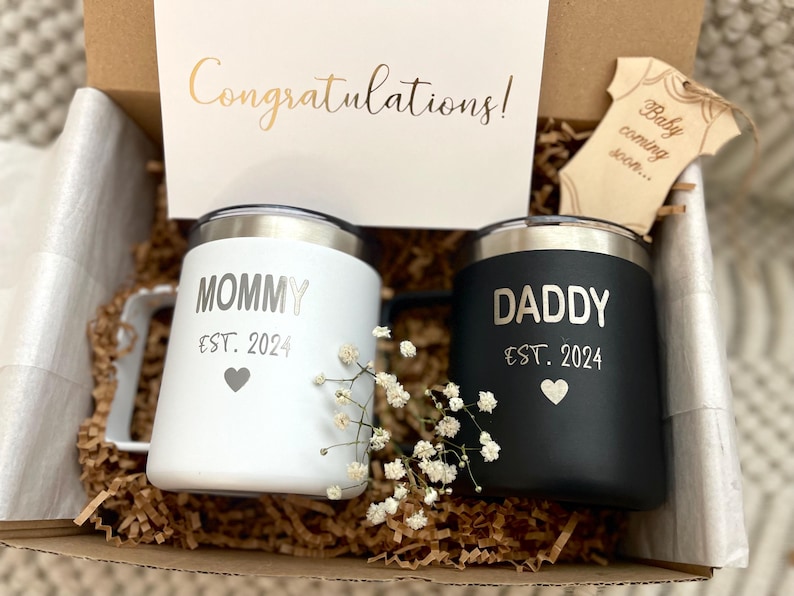 Puede incluir: Una caja de regalo con dos tazas aisladas, una blanca y otra negra, con bordes plateados. La taza blanca dice "MOMMY EST. 2024" y la taza negra dice "DADDY EST. 2024". Una tarjeta dice "&iexcl;Enhorabuena!" y se incluye una etiqueta de madera con forma de body de beb&eacute;.