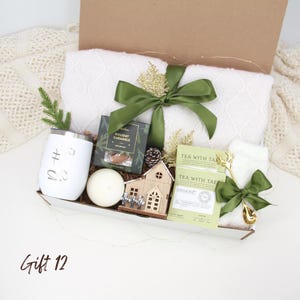 Puede incluir: Una caja de regalo llena de una manta blanca, un vaso blanco, una vela, paquetes de t&eacute;, caramelos y una casa de madera decorativa. La caja est&aacute; atada con una cinta verde. El texto "Gift 12" est&aacute; en la parte inferior izquierda.