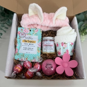 May include: A gift box filled with self-care items. Includes a pink headband, face mask, sugar scrub, bath bomb, hair clip, and wrapped candies. The box is lined with brown paper and has a white interior.