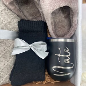 Pode incluir: Uma caixa de presente cheia de itens aconchegantes, incluindo um par de pantufas cinzas felpudas, meias pretas, um cobertor de malha branco e cinza e um copo de a&ccedil;o inoxid&aacute;vel preto com a palavra "Nate" gravada.