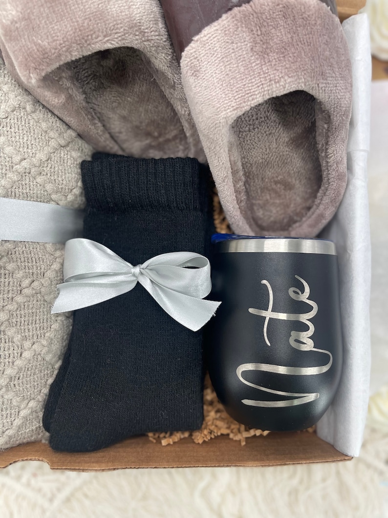 Pode incluir: Uma caixa de presente cheia de itens aconchegantes, incluindo um par de pantufas cinzas felpudas, meias pretas, um cobertor de malha branco e cinza e um copo de a&ccedil;o inoxid&aacute;vel preto com a palavra "Nate" gravada.