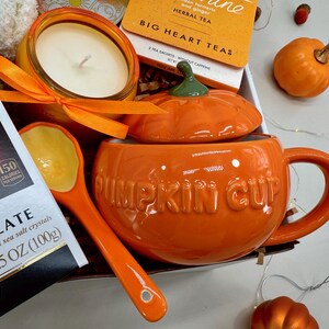 May include: An orange ceramic pumpkin-shaped mug with a lid, labeled "PUMPKIN CUP", is the focal point of this image. A matching spoon, candle, tea, and chocolate are also present, creating a fall-themed gift set. The chocolate is 3.5 OZ (100g).