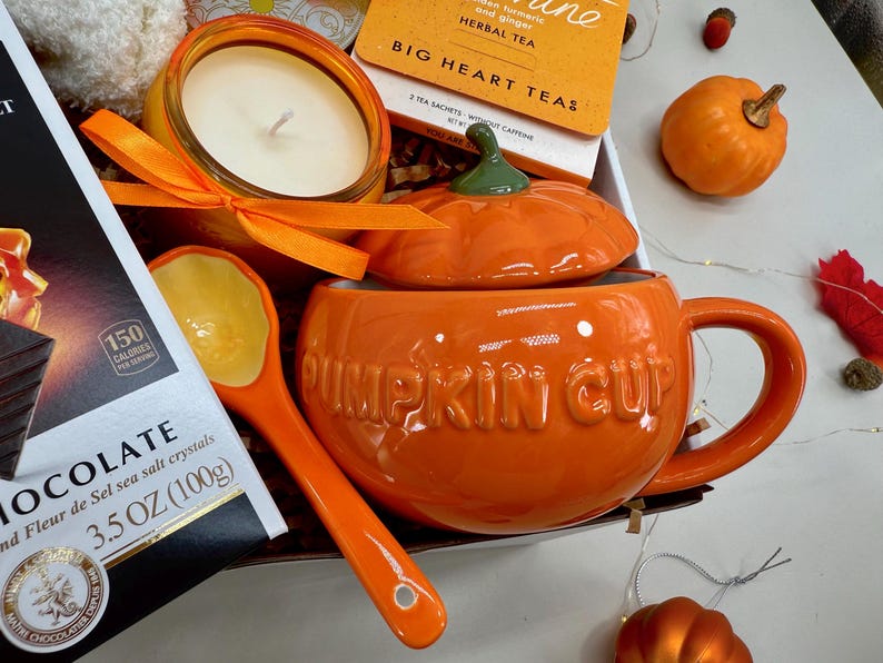 May include: An orange ceramic pumpkin-shaped mug with a lid, labeled "PUMPKIN CUP", is the focal point of this image. A matching spoon, candle, tea, and chocolate are also present, creating a fall-themed gift set. The chocolate is 3.5 OZ (100g).