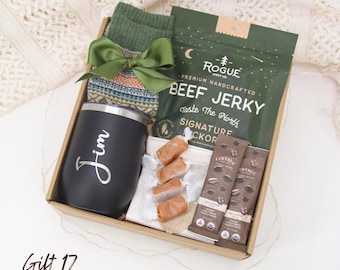 May include: A gift box containing a black tumbler with "Jim" in script, green socks, a green bow, beef jerky, coffee bars, and wrapped candies. The box is brown and sits on a white surface.