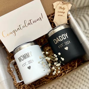 Puede incluir: Una caja de regalo con dos tazas aisladas, una blanca y otra negra, con las palabras "Mommy" y "Daddy" y "Est. 2024". Se incluye una tarjeta con "&iexcl;Enhorabuena!" y un recorte de ropa de beb&eacute; de madera.