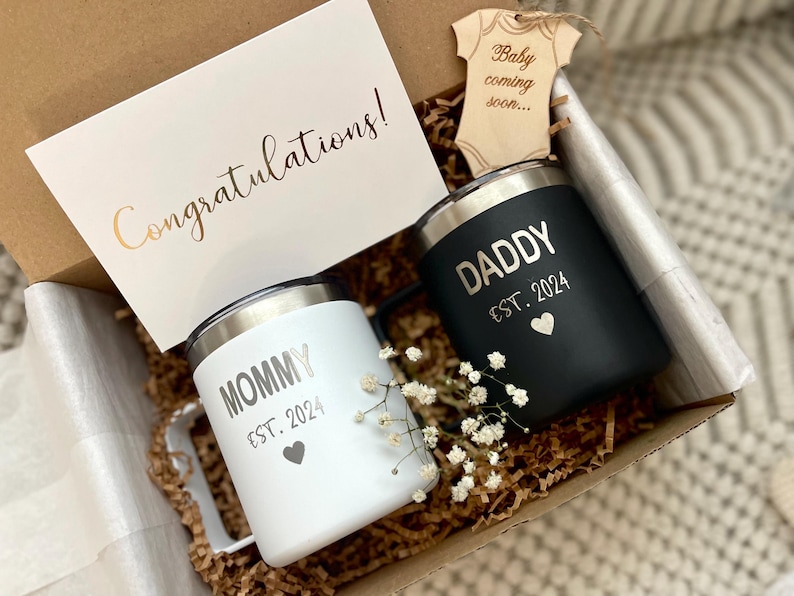 Puede incluir: Una caja de regalo con dos tazas aisladas, una blanca y otra negra, con las palabras "Mommy" y "Daddy" y "Est. 2024". Se incluye una tarjeta con "&iexcl;Enhorabuena!" y un recorte de ropa de beb&eacute; de madera.