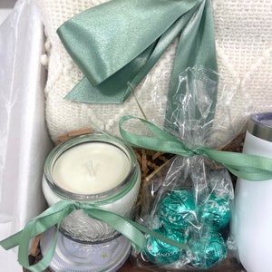 May include: A gift basket with a white blanket, a candle in a glass jar, a bag of green chocolate truffles, and a white mug.