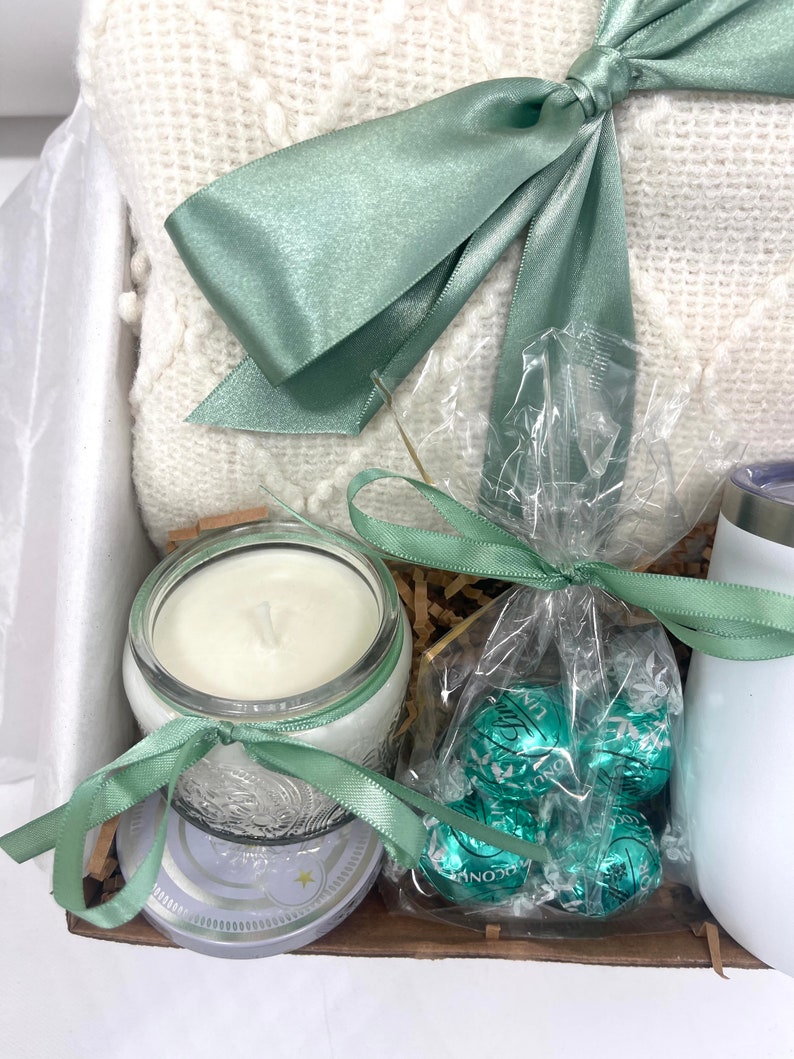 May include: A gift basket with a white blanket, a candle in a glass jar, a bag of green chocolate truffles, and a white mug.