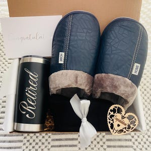 Peut inclure: Une bo&icirc;te-cadeau remplie d'un gobelet isotherme noir et argent avec le mot "Retrait&eacute;" dessus, une paire de pantoufles bleues avec une doublure en polaire grise, une couverture ray&eacute;e noir et blanc et un c&oelig;ur en bois avec le mot "F&eacute;licitations" dessus.