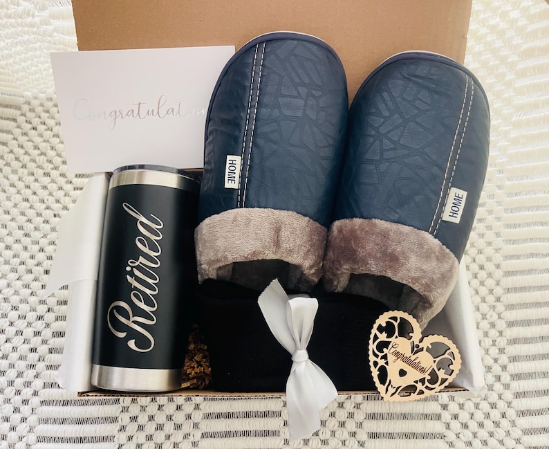 Peut inclure: Une bo&icirc;te-cadeau remplie d'un gobelet isotherme noir et argent avec le mot "Retrait&eacute;" dessus, une paire de pantoufles bleues avec une doublure en polaire grise, une couverture ray&eacute;e noir et blanc et un c&oelig;ur en bois avec le mot "F&eacute;licitations" dessus.