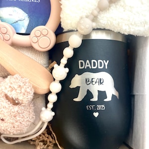 Może przedstawiać: Czarny kubek z napisem "DADDY BEAR" i białą sylwetką niedźwiedzia. Na kubku znajduje się r&oacute;wnież napis "EST. 2025" i serce. Obraz zawiera zestaw prezentowy dla dziecka z klipsem do smoczka, książką i pluszową zabawką.