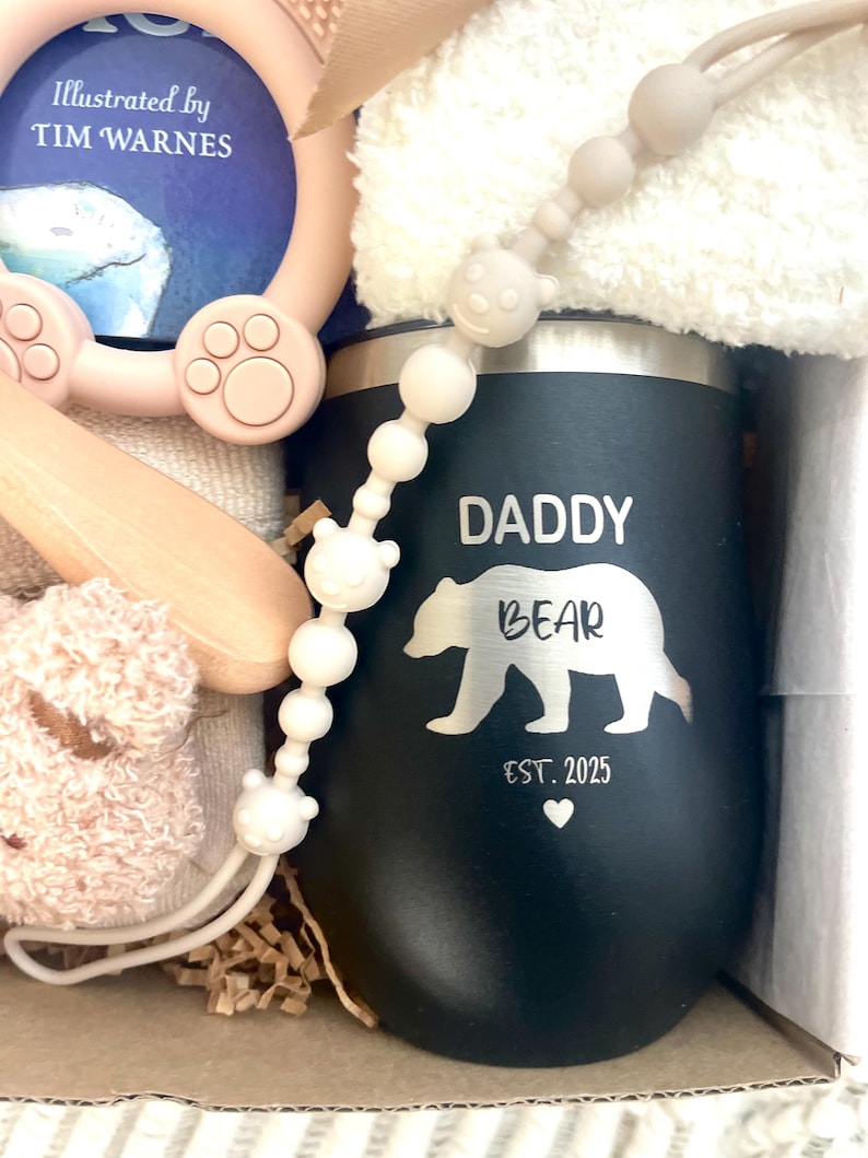 Może przedstawiać: Czarny kubek z napisem "DADDY BEAR" i białą sylwetką niedźwiedzia. Na kubku znajduje się r&oacute;wnież napis "EST. 2025" i serce. Obraz zawiera zestaw prezentowy dla dziecka z klipsem do smoczka, książką i pluszową zabawką.