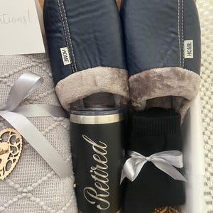 Peut inclure: Un panier-cadeau rempli d'une couverture grise, d'une tasse de voyage noire et argent&eacute;e avec le mot "Retrait&eacute;" dessus, d'une paire de chaussettes noires et d'une paire de pantoufles bleu marine avec le mot "HOME" dessus.