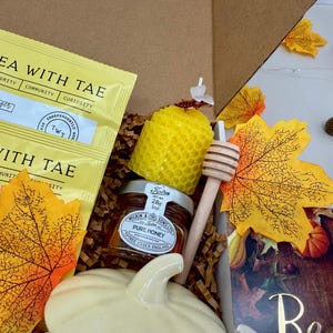 May include: A gift box with tea packets, a jar of honey, a honey dipper, a yellow honeycomb candle, and a white ceramic pumpkin. The box also contains decorative fall leaves and small pumpkins. The tea packets read "Tea with Tae".