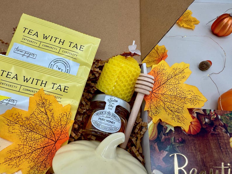 May include: A gift box with tea packets, a jar of honey, a honey dipper, a yellow honeycomb candle, and a white ceramic pumpkin. The box also contains decorative fall leaves and small pumpkins. The tea packets read "Tea with Tae".