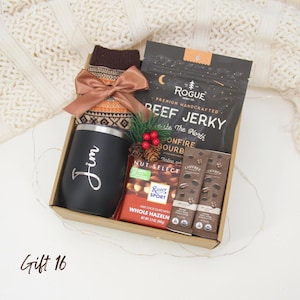 Puede incluir: Caja de regalo con un vaso negro con "Jim", carne seca, barras de chocolate, calcetines estampados y un lazo. La caja tambi&eacute;n contiene una ramita de vegetaci&oacute;n y una barra de chocolate. El texto "Gift 16" est&aacute; en la parte inferior.